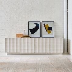 two framed art pieces sitting on top of a white shelf next to a brick wall