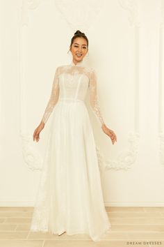 a woman standing in front of a white wall wearing a wedding dress with long sleeves