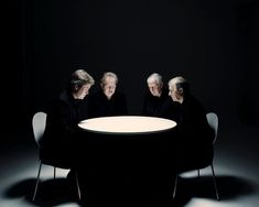 three people sitting at a round table in the middle of a dark room with one person looking on