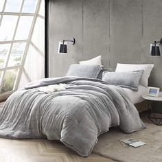 a bed with grey linens and pillows in a bedroom next to an open window