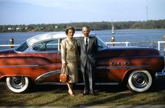 a man and woman standing next to an old fashioned car in front of the water