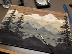 a piece of wood that has been painted with mountains and trees on it, along with paintbrushes