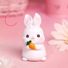 a small white bunny holding a carrot in its hand and sitting next to some daisies