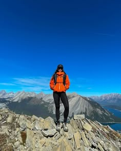 a man in an orange jacket standing on top of a rock formation with mountains in the background