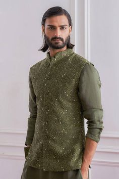 a man with long hair and beard wearing a green shirt standing in front of a white wall