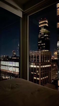 a bed sitting in front of a window next to a night time cityscape