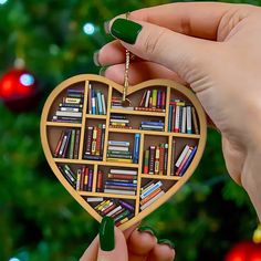 two hands holding a heart shaped book shelf ornament in front of a christmas tree