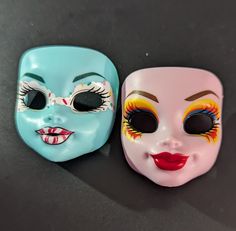 two plastic masks with painted faces on them sitting next to each other in front of a black surface