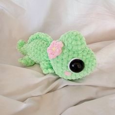 a small green stuffed animal laying on top of a white bed sheet with a pink flower