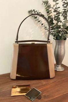 "1950s 1960s Leon Of California Color Block Two Tone Brown Vinyl Top Handle Handbag, Vintage Purse, Mid Century Accessory  Chic 1950s 1960s top handle two tone brown vinyl handbag made by Leon Of California that features: The front and sides of the handbag is a chocolate brown and cappuccino colored vinyl. The back of the bag is a solid chocolate brown vinyl. It has a chocolate brown single handle strap connected to a decorative brown bar. There is gold tone metal hardware with a thumb lift closure that works well and closes tightly. Opens to a deep burgundy satin lining with one interior side open pocket with gold trim. Stamped Leon Of California. comes with a gold metal mirror case with the two side mirror included. Beautiful vintage condition *the metal hardware is bright and shiny.  Th Mid-century Brown Evening Bags, Mid-century Brown Shoulder Bag, 1960s Handbags, Mid-century Brown Leather Bags, Mid Century Accessories, California Colors, 1960 Handbags Purses, Deep Burgundy, Top Handle Handbags