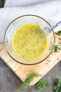 a whisk in a glass bowl on top of a cutting board with herbs