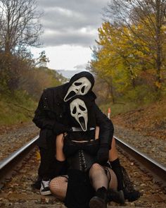 two people sitting on train tracks with masks covering their faces and mouths, one person is holding the other's head