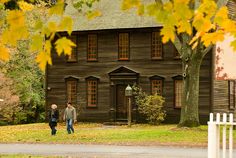 two people walking in front of an old wooden house with fall leaves on the ground