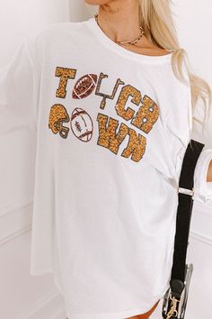 - Football, but make it fabulous with this shimmering tee! Its unique sequined design makes it a perfect addition to any sporty fashionista's game day collection. Stay at the top of the tailgate game with this sporty-chic piece! - Unlined 100% cotton material - A sequined white, black, gold, and brown hued 'TOUCHDOWN' design featuring footballs, a goal post, and a helmet - A crew cut neckline - Short, loose sleeves - A relaxed silhouette that ends in a straight hemline - This piece is perfect fo White Team Spirit T-shirt For Fall, White Tops With Team Spirit For Fall, White Sports Fan Top For Fall, White Tops For Fall With Team Spirit Style, White Tops For Fall Sports Fans, White Oversized T-shirt For Sports Events, Oversized White T-shirt For Sports Events, Collegiate White T-shirt For Fall, White Collegiate T-shirt For Fall