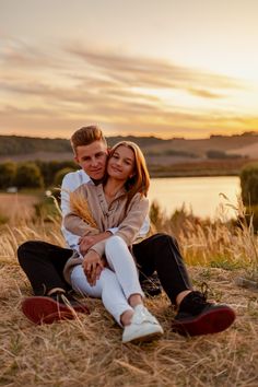 a man and woman sitting on the ground in front of a body of water at sunset