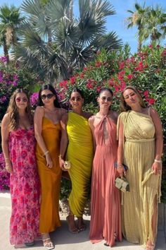 four women in long dresses posing for a photo together with palm trees and flowers behind them