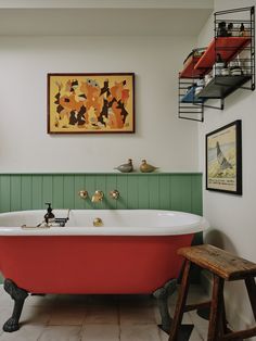 a red bath tub sitting in a bathroom next to a wooden stool and painting on the wall