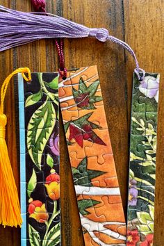 three colorful bookmarks with tassels hanging from them on a wooden table next to other books