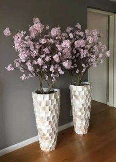 two white vases with pink flowers in them on a wooden floor next to a gray wall