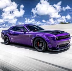 a purple sports car driving down the road with clouds in the sky behind it on a sunny day