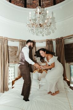 a man standing next to a woman on top of a bed in front of a chandelier