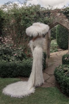 a woman in a long white dress standing next to a stone wall and shrubbery