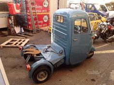 an old blue truck is parked in a lot next to other motorcycles and scooters