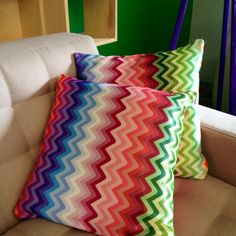 two colorful pillows sitting on top of a couch next to a white chair and green wall