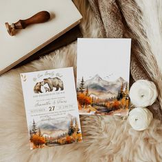 two wedding stationery cards with bears and mountains on them sitting on a fur rug