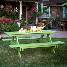 a green picnic table with fruit on it in front of a brick building and flowers