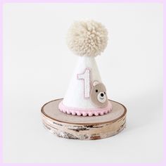 Party hat is made of - white merino felt - pink pompom trim - elastic band - beige pom - white/pink layered birthday number - bear face Size is  H 12 cm W 8 cn Cute White Hat For First Birthday, Cute White Hat For Birthday, White Birthday Party Hat Supplies, Playful White Party Supplies For First Birthday, Whimsical White Birthday Party Supplies, Whimsical White Party Supplies For Birthday, Playful White Hat For First Birthday, Playful White Hat For Birthday, Playful White Hats For Birthday