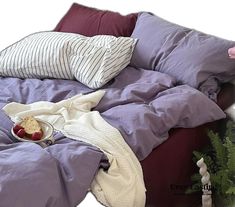 an unmade bed with purple sheets, pillows and fruit on the bottom shelf next to it