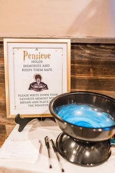 a table topped with a metal bowl filled with blue liquid next to a sign that reads pensite