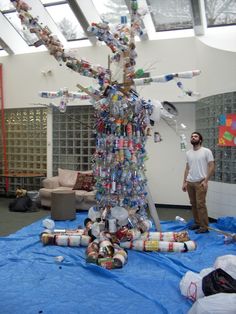 a man standing next to a large tree made out of cans and plastic bottle caps