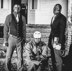 three men standing on the front steps of a house, one wearing a suit and tie