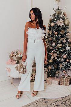 a woman standing in front of a christmas tree wearing white jumpsuits and holding a purse