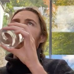 a woman drinking water from a glass in front of a window with multiple pictures behind her