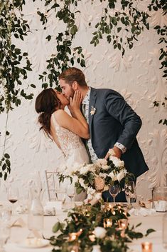 a bride and groom kissing in front of greenery at the head table with candles