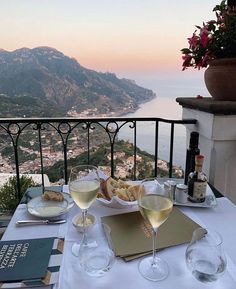 two glasses of wine are sitting on a table overlooking the water and mountains in italy