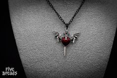 Add a touch of gothic romance to your jewelry collection with our Silver Winged Sword Pendant Necklace. This unique piece combines dark elegance with intricate design, making it a perfect accessory for those who appreciate the beauty of gothic style. Pendant Details: Design: A silver-colored zinc alloy pendant featuring a winged sword penetrating a red enamel heart with a tiny skull inside. Material: High-quality zinc alloy metal (Lead-Nickel Free). Chain Details: Length: 18-19 inches. Color: Bl Gothic Heart Pendant Jewelry, Gothic Jewelry For Cosplay On Valentine's Day, Red Heart Pendant Necklace, Red Heart Pendant, Necklace Gothic, Gothic Romance, Red Pendants, Silver Wings, China Jewelry