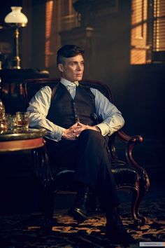 a man sitting in a chair wearing a vest and tie with his hands folded out