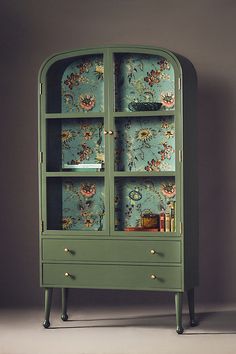 a green cabinet with floral wallpaper on the back and shelves below it, against a gray background