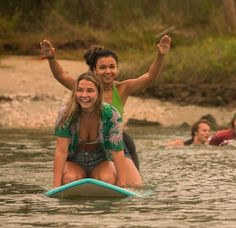 two girls are riding on a surfboard in the water while another girl smiles and waves