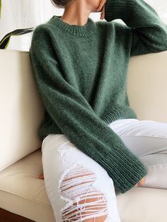 a woman sitting on top of a white couch wearing a green sweater and ripped jeans