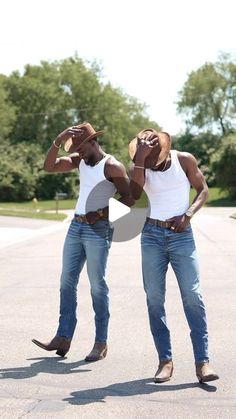 two men in jeans and cowboy hats are standing on the street with their hands behind their heads