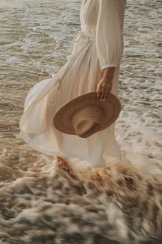 a woman in a white dress and hat walking into the ocean