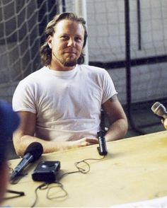 a man sitting at a table with microphones in front of him and another person standing next to him