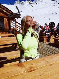 a woman drinking water from a bottle while sitting at a wooden bench