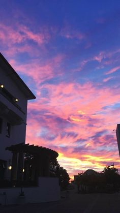 the sun is setting over some buildings and trees in front of it with purple clouds