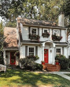 a white house with red front door and window boxes on the windowsills is surrounded by greenery